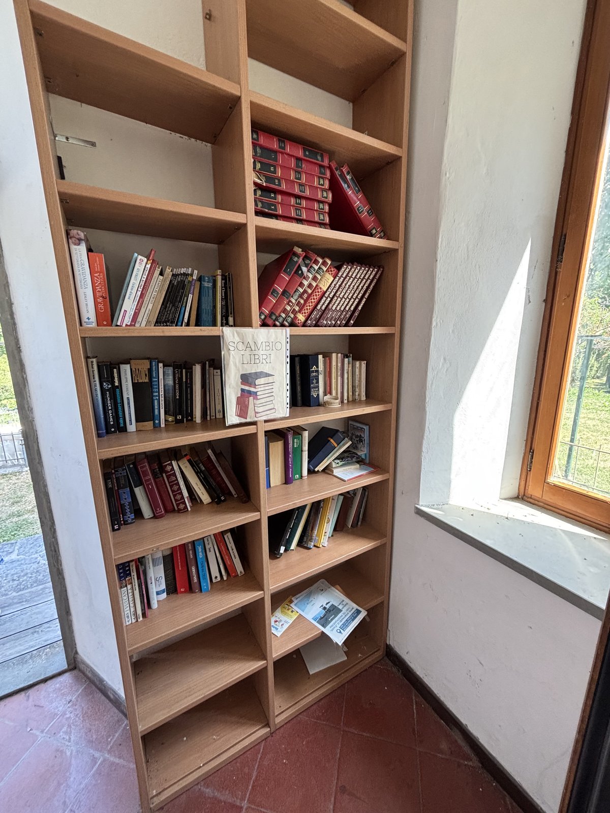 A tall wooden book exchange shelf stands indoors beside a sunlit window, with several shelves filled with assorted books and magazines. A small sign reading “Scambio Libri” marks it as a community book swap, and the simple corner setting gives it a cozy, informal feel.