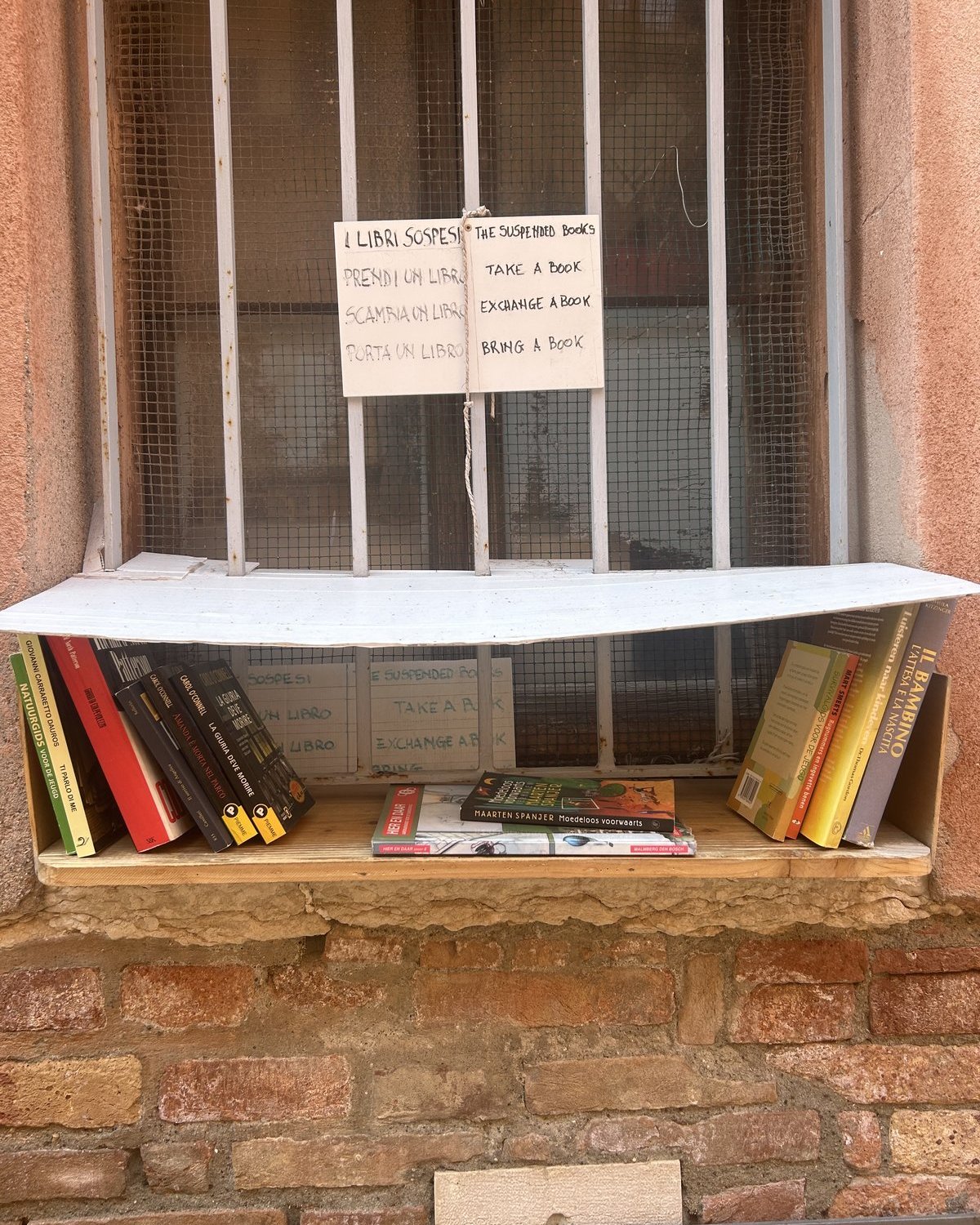 A small free book exchange is built into a barred window set in a weathered brick wall in Venice. A handwritten bilingual sign says “The Suspended Books” and invites visitors to take, exchange, or bring a book, with several paperbacks lined up on the shelf below.