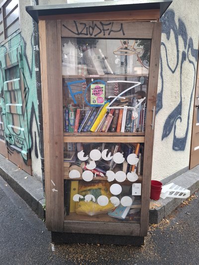 A tall wooden, glass-fronted book exchange cabinet stands against a graffiti-covered urban wall at Hallwylplatz in Zurich. It is densely packed with books in several languages, with colorful posters, stickers, and painted markings covering the doors and surrounding facade.