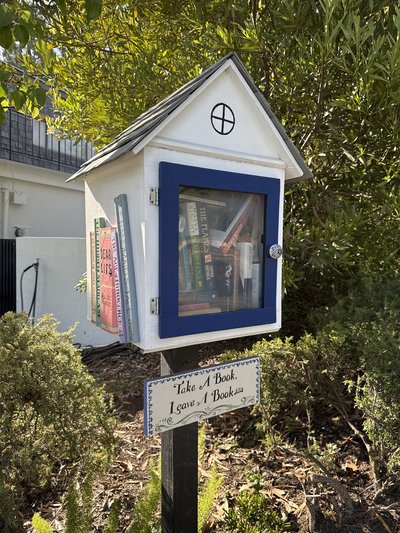 A charming white, cottage-style book exchange with a steep shingled roof, blue-trimmed glass door, and books displayed inside and along the side. It stands on a dark wooden post amid leafy shrubs, with a decorative sign inviting visitors to take and leave a book.