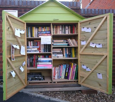 A large lime-green wooden book exchange stands against a brick wall, with both doors open to reveal several shelves packed with books. Handwritten notes and messages are clipped to strings across the doors, including a notice that games and DVDs are not accepted.